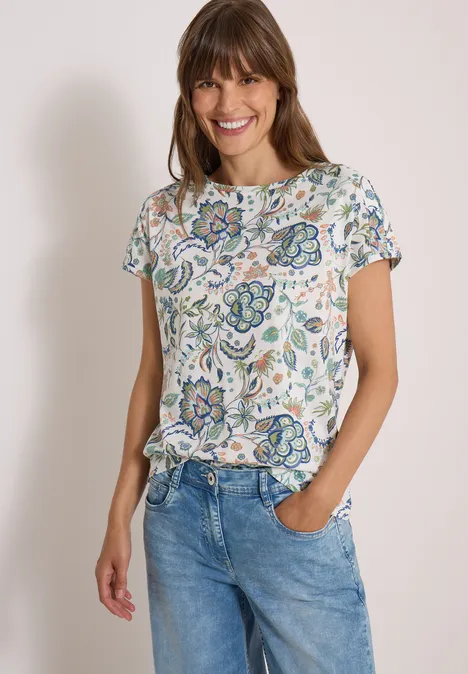 Een lachende vrouw draagt een kleurrijk bloemenblouse en een blauwe spijkerbroek.