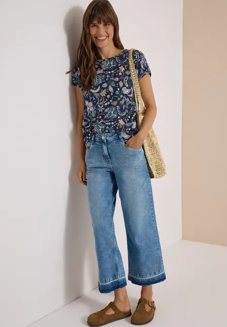 Vrouw met bloemendessin top, wijde cropped jeans, strooien tas en bruine clogs, glimlachend tegen een witte muur.