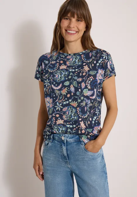 Lachende vrouw in blauwe bloementop en lichte jeans, ontspannen pose met één hand in zak.