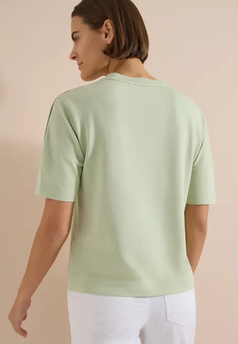 Femme de dos portant un haut manches courtes vert clair et un pantalon blanc.