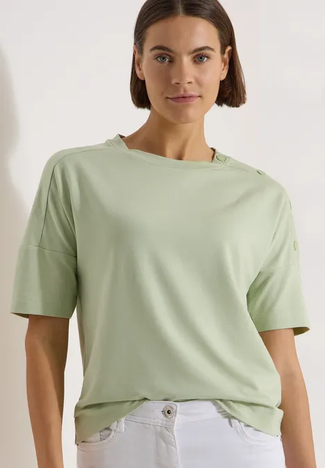 Femme porte un t-shirt vert clair et un jean blanc sur fond blanc.