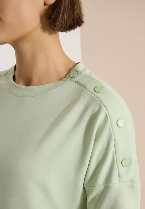 Gros plan sur un haut vert pâle à col rond avec des boutons décoratifs épaule.