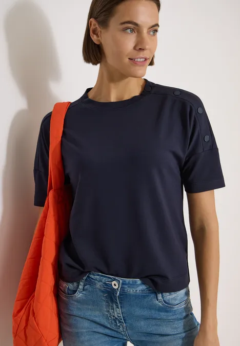 Vrouw in donkerblauw T-shirt met schouderknopen, spijkerbroek en rode tas bij lichte muur.