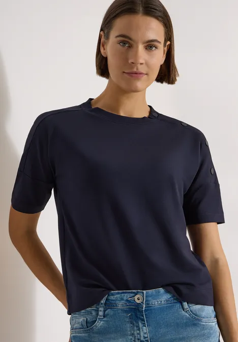 Een persoon in een donkerblauw T-shirt en spijkerbroek tegen een lichte achtergrond.