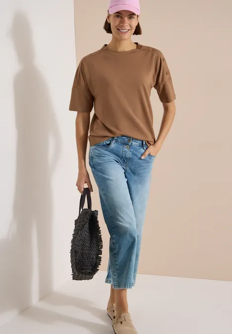 Personne en t-shirt marron, jean bleu et sac noir sur fond clair.