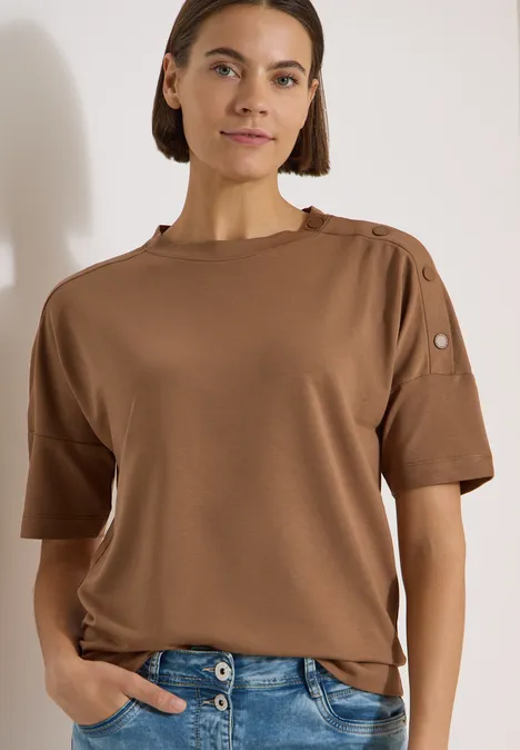 Personne portant un t-shirt marron à manches courtes avec boutons à l'épaule.