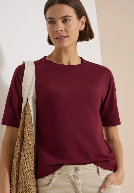 Femme portant un T-shirt bordeaux, un pantalon beige et un sac épaule beige devant un fond neutre.