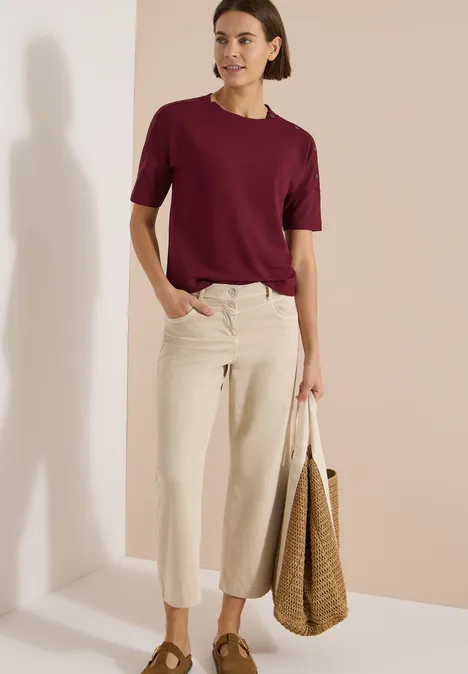 Femme en t-shirt bordeaux, jean large clair et sac en osier devant un fond neutre.