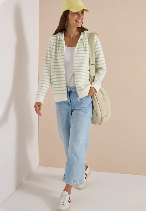 Frau trägt gestreifte Kapuzenjacke, weißes Top, weite Jeans, weiße Sneaker, gelbe Mütze und Tasche.