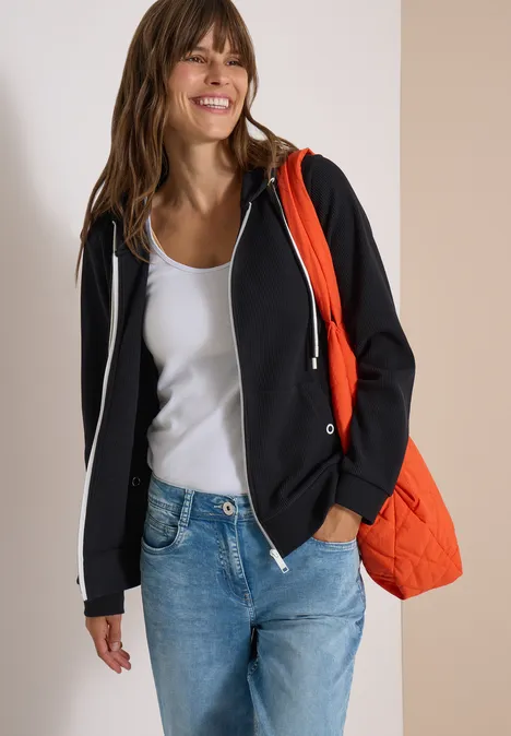 Een glimlachende vrouw in wit topje, zwart vest met capuchon, spijkerbroek en oranje tas.