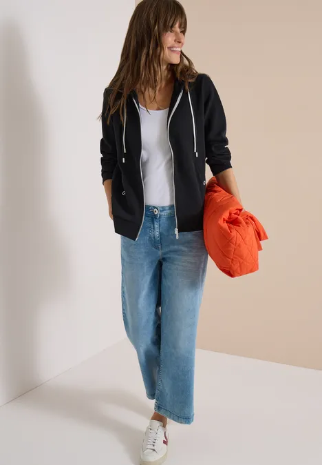 Frau in schwarzer Jacke, weißem Top und blauen Jeans trägt weiße Sneaker und hält eine orangefarbene Tasche.