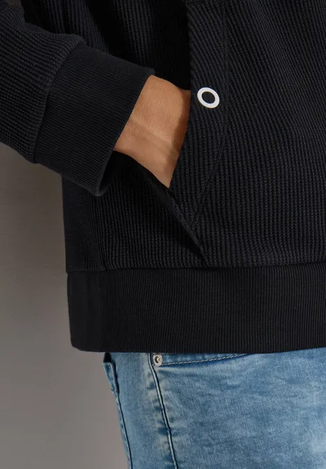 Person trägt schwarzen Hoodie mit Hand in der Tasche und blaue Jeans.