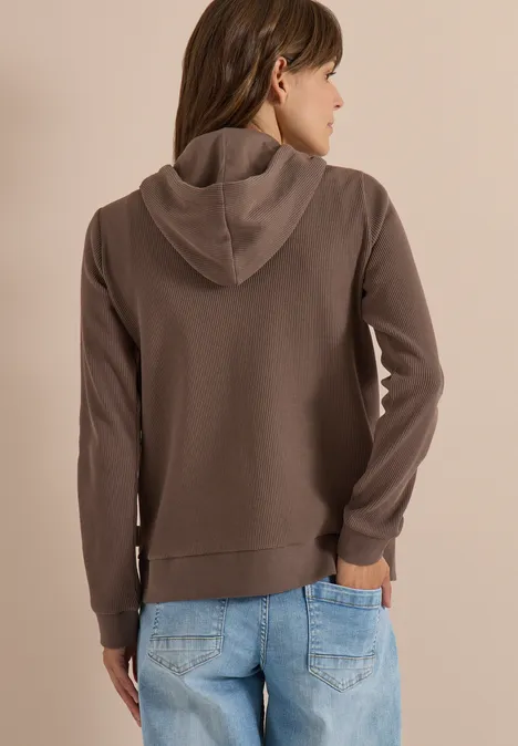 Frau von hinten mit braunem Kapuzenpullover und blauen Jeans vor hellem Hintergrund.