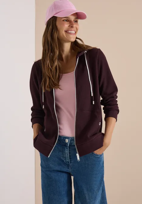 Een lachende vrouw draagt een roze pet, een bordeaux hoodie, een roze top en een blauwe jeans.