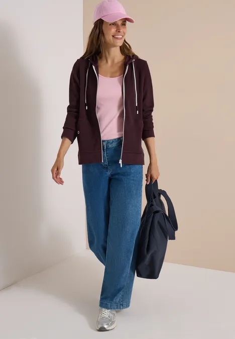 Frau in pinkem Top, violetter Kapuzenjacke, weiter Jeans und Silber-Sneakern, rosafarbener Cap, sie hält eine blaue Jacke.