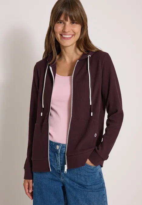 Junge Frau lächelt in einem dunkelvioletten Hoodie, rosa Top und Jeans vor weißer Wand.