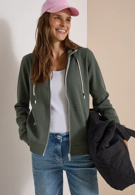 Vrouw draagt groene capuchonsweater, wit topje, jeans en roze pet, houdt een zwarte jas vast, glimlacht.
