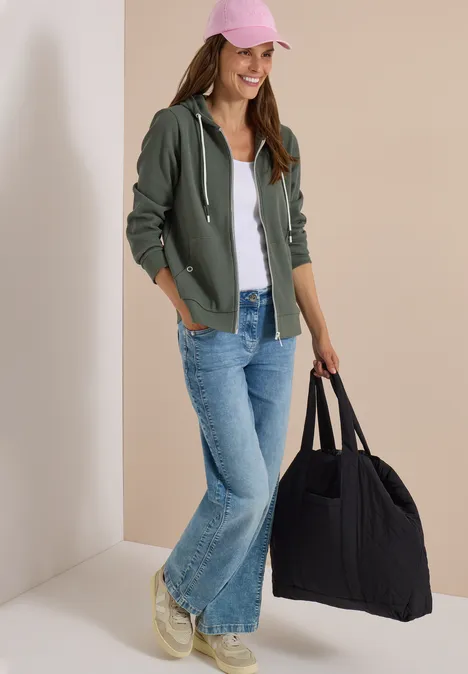 Junge Frau in Jeans, Hoodie und rosa Cap, die eine große schwarze Tasche trägt.