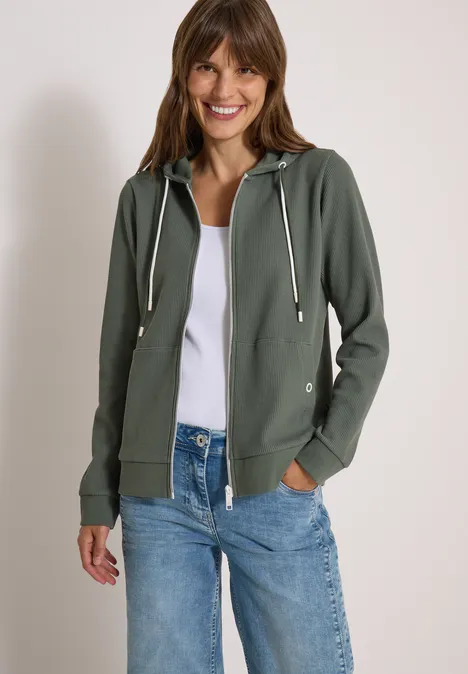 Lächelnde Frau in grüner Kapuzenjacke und Jeans vor hellem Hintergrund.
