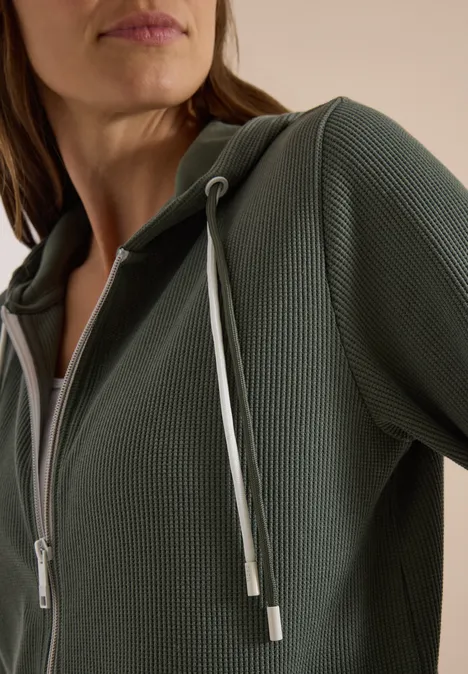 Nahaufnahme einer Person in einem olivgrünen, feinstrukturigen Hoodie mit Reißverschluss und hellem Kordelzug.