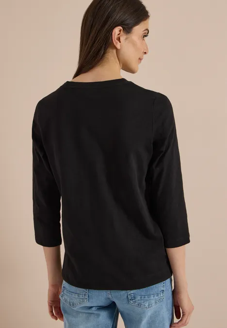 Rückansicht einer Frau mit schwarzem Dreiviertelarm-Shirt und Jeans vor hellem Hintergrund.