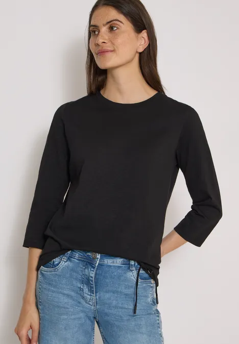 Eine Frau in schwarzem 3/4-Arm-Shirt und blauen Jeans steht locker vor weißem Hintergrund.