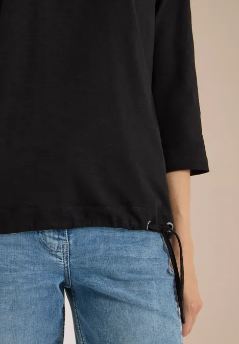 Person trägt blaue Jeans und schwarzen Dreiviertelärmel-Pullover mit seitlicher Kordel, vor beigem Hintergrund.