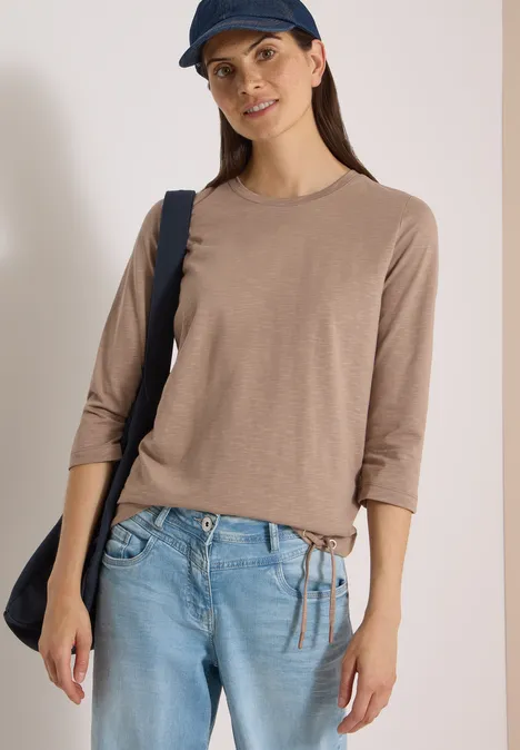 Frau in beige 3/4-Arm-Shirt, hellblauen Jeans und dunkler Umhängetasche.