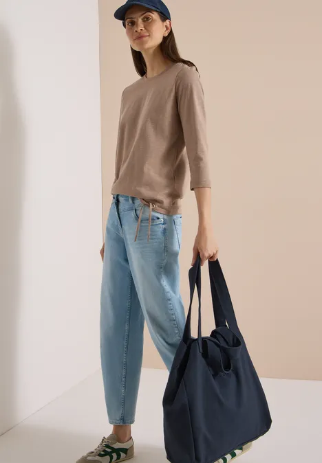 Frau in lässigem Outfit mit beiger Bluse, hellblauen Jeans und Cap hält eine große dunkelblaue Tasche.