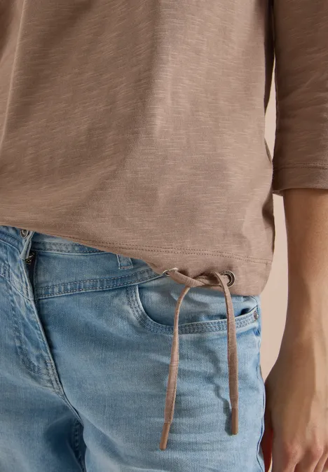 Nahaufnahme eines Oberkörpers mit beigem Shirt und blauen Jeans mit seitlichen Bändern am Bund.