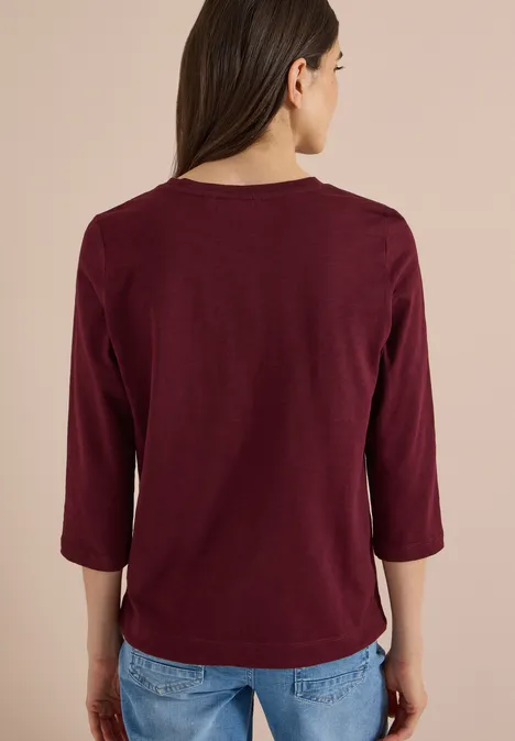 Rückansicht einer Person in einem bordeauxroten Dreiviertelarmshirt und blauen Jeans vor neutralem Hintergrund.
