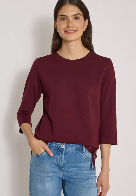 Dame in blauem Jeans und burgunderrotem Langarm-Shirt, das an der Taille gebunden ist.
