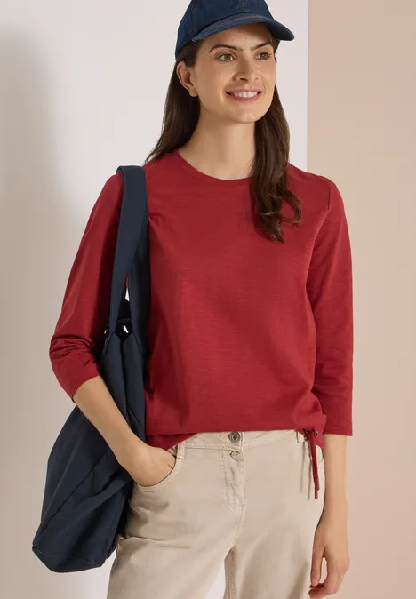 Jonge vrouw met rood shirt, beige broek, blauwe pet en blauwe tas, glimlachend.