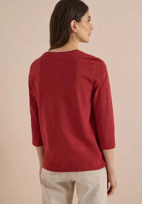 Frau in rotem 3/4-Arm-Shirt, Rückenansicht, trägt beige Hose.