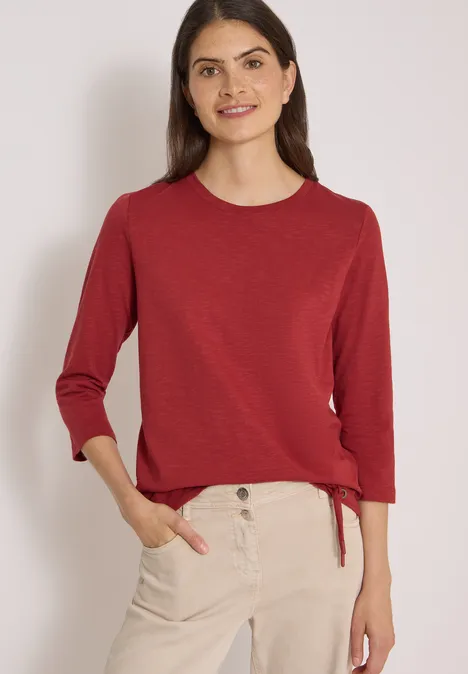 Frau trägt roten Dreiviertelarm-Pullover und helle Chinohose, steht lächelnd mit Händen in den Taschen.
