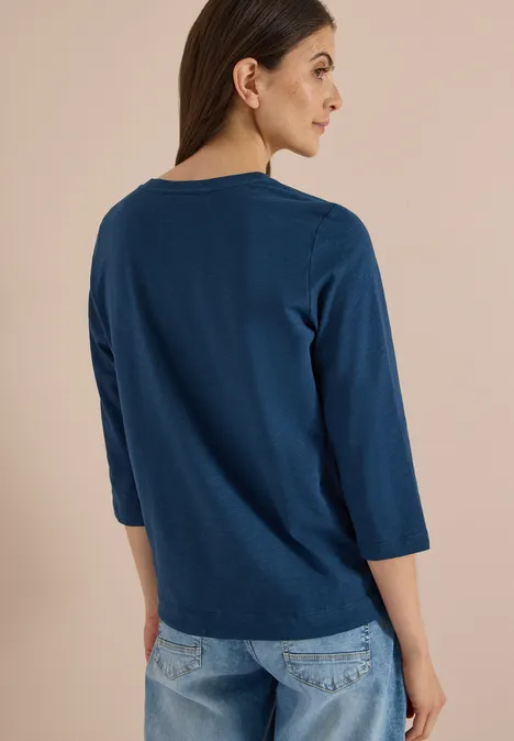 Eine Frau in Jeans und dunkelblauer 3/4-Arm-Bluse mit Rundhalsausschnitt, seitlich von hinten fotografiert.