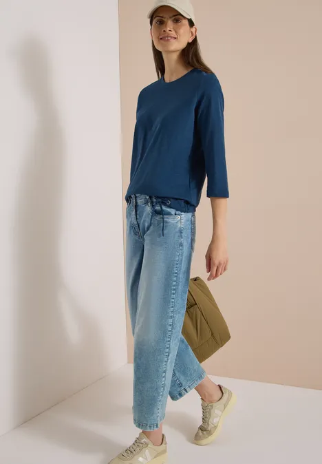 Frau in blauem Shirt, weit geschnittenen Jeans, Sneakern und Cap, trägt eine grüne Tasche.