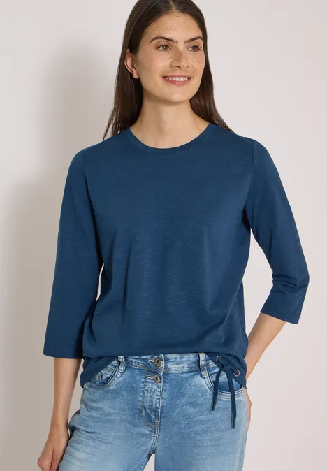 Frau trägt marineblaues 3/4-Arm-Shirt mit seitlicher Schleife und helle Jeans, lächelnd vor hellem Hintergrund.