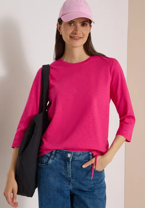 Junge Frau in pinkem Pullover und Cap, blauer Jeans, mit schwarzer Tasche auf der Schulter.