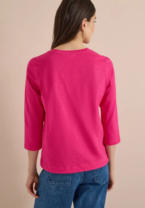 Frau von hinten in pinkem 3/4-Arm-Shirt und blauen Jeans vor hellem Hintergrund.