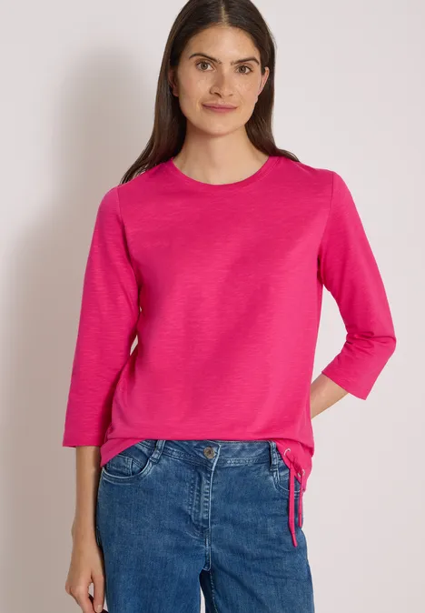Frau in pinkem Langarmshirt und Jeans, frontal stehend vor hellem Hintergrund
