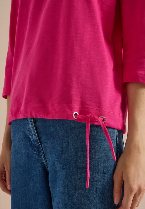 Eine Person trägt ein pinkes Shirt mit seitlichen Kordelzügen und blaue Jeans.