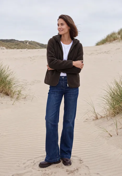 Vrouw in jeans en jas staat ontspannen met gekruiste armen op duinen bij het strand.