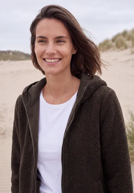 Een glimlachende vrouw in een fleece trui staat op een strand met duinen op de achtergrond.