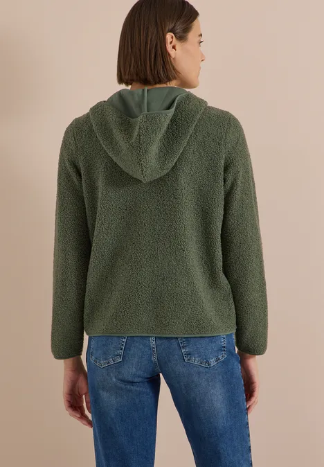 Frau von hinten in grüner Teddy-Fleecejacke mit Kapuze und blauen Jeans vor beigem Hintergrund.