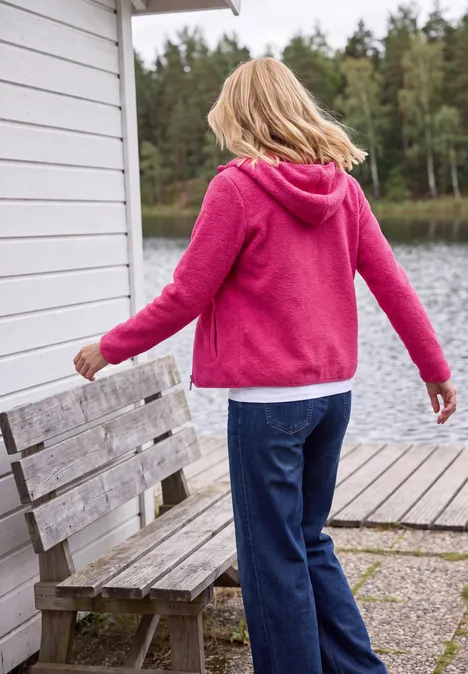 Une personne en sweat rose et jean se tient sur un ponton au bord d’un lac, près d’un banc en bois et d’une forêt.