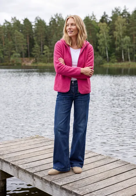 Une femme en pull rose et jean se tient détendue sur un ponton en bois au bord d’un lac, forêt en arrière-plan.