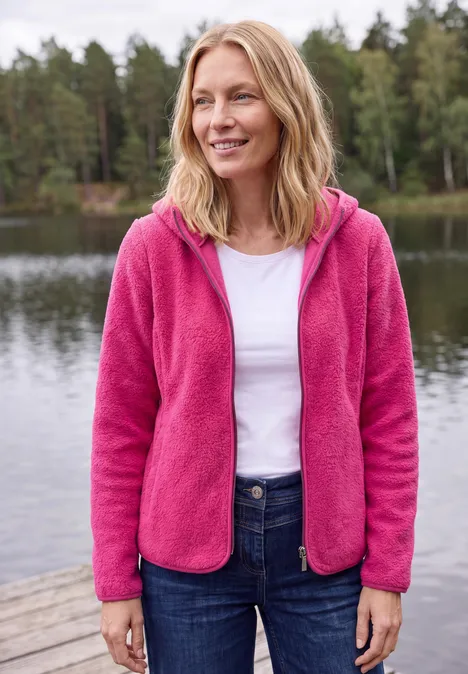 Femme debout sur une jetée au bord d’un lac, veste polaire rose, chemise blanche et jean.