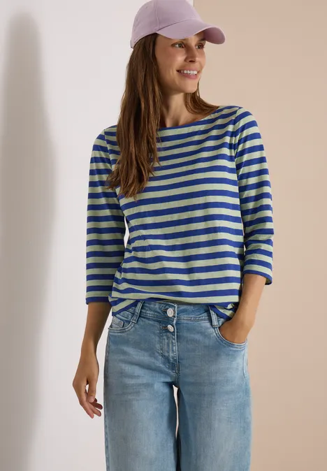 Vrouw in gestreept shirt, spijkerbroek en roze pet, glimlachend met één hand in haar zak.
