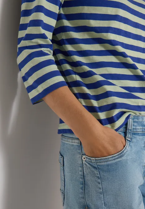 Person trägt blau-weiß gestreiftes 3/4-Ärmel-Shirt und Jeans, Hand steckt in Hosentasche.